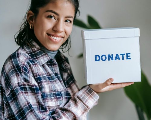 Happy ethnic lady in checkered shirt and bun looking at camera and smiling while demonstrating donation box on background with potted plant near white wall